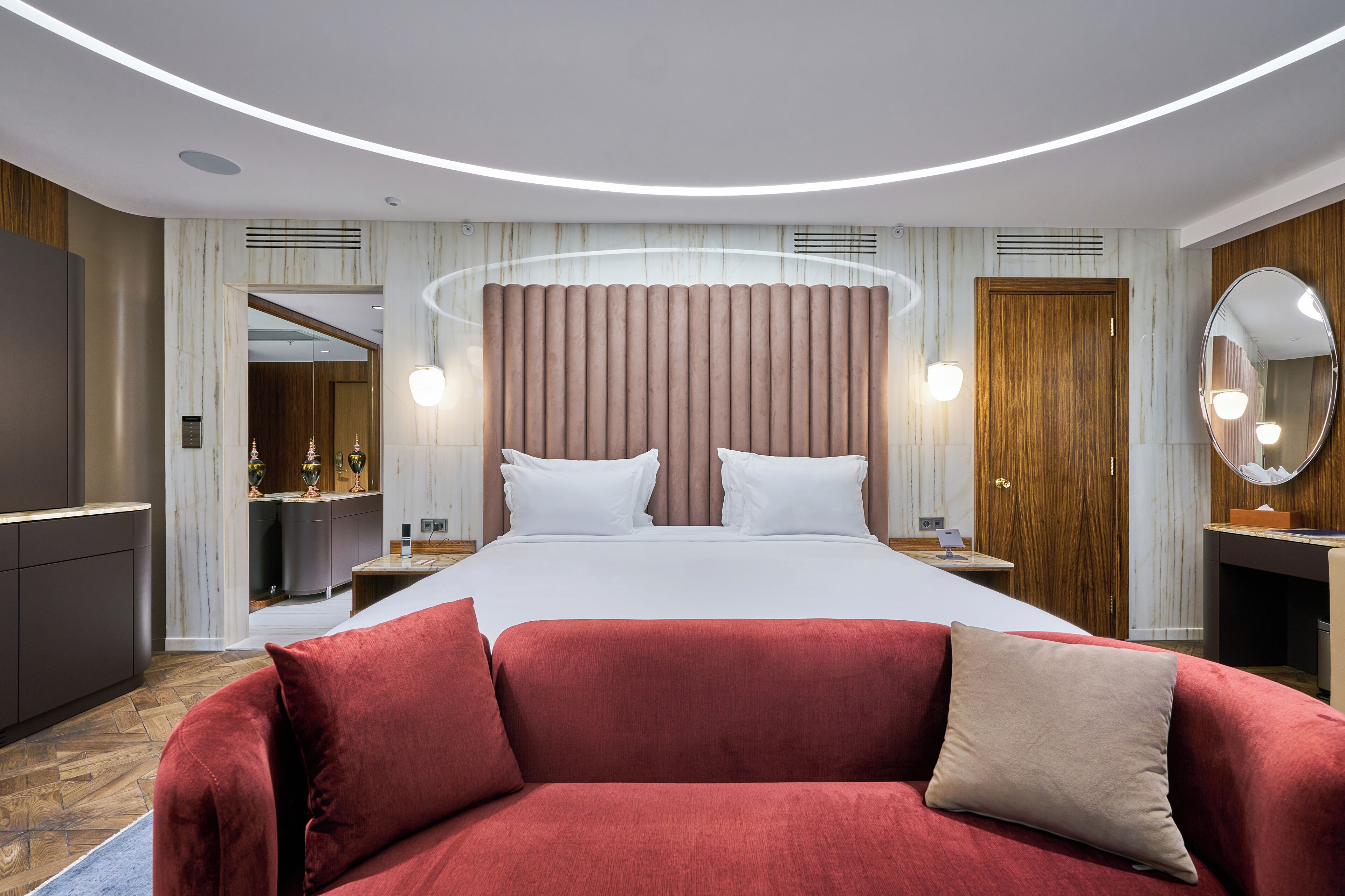 A modern hotel room with a large bed, a red sofa, a wooden door, and a mirror.