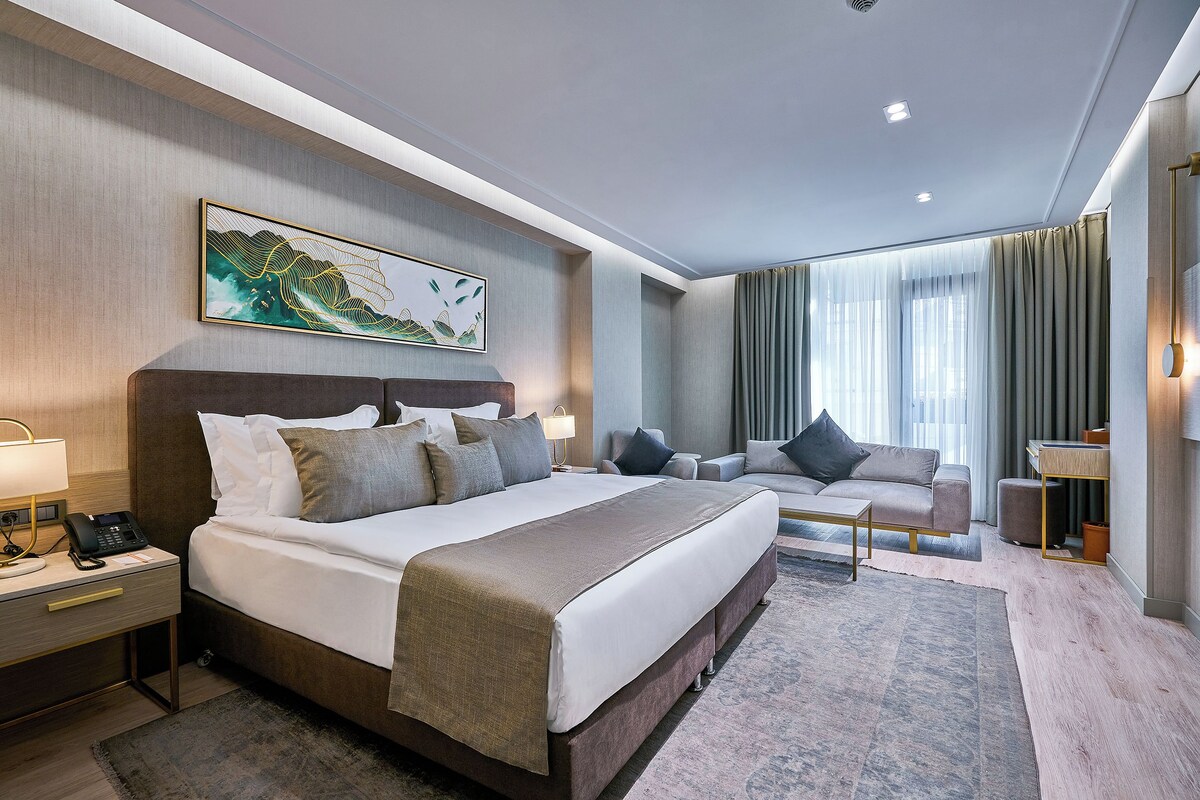 A modern hotel room with a large bed, a desk with a lamp, a sofa, and a large window with curtains.