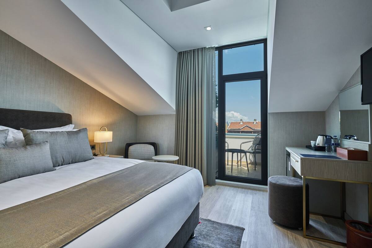 A modern hotel room with a large bed, a desk, a chair, and a balcony with a view.