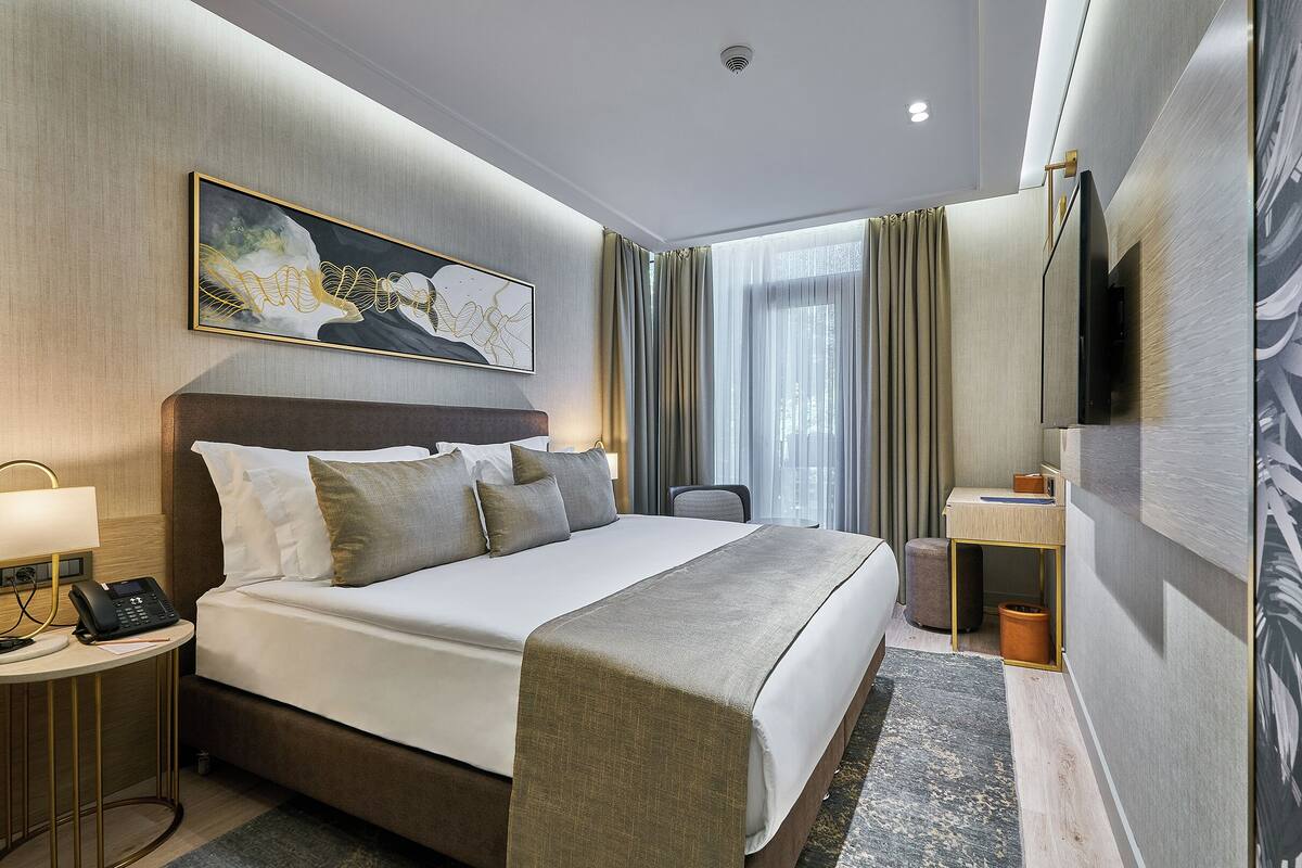 A hotel room with a bed, bedside table, lamp, phone, and a painting on the wall.