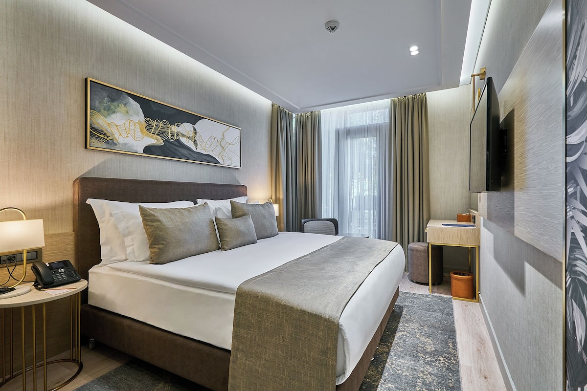 A hotel room with a bed, bedside table, lamp, phone, and a painting on the wall.