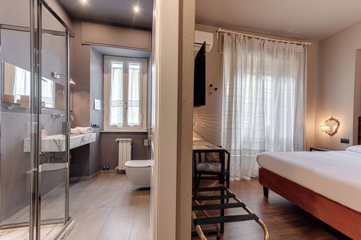 Ein modernes Hotelzimmer mit Duschkabine aus Glas, einem separaten Badezimmer, einem Bett mit Holz Kopfteil und einem an der Wand befestigten Fernseher.