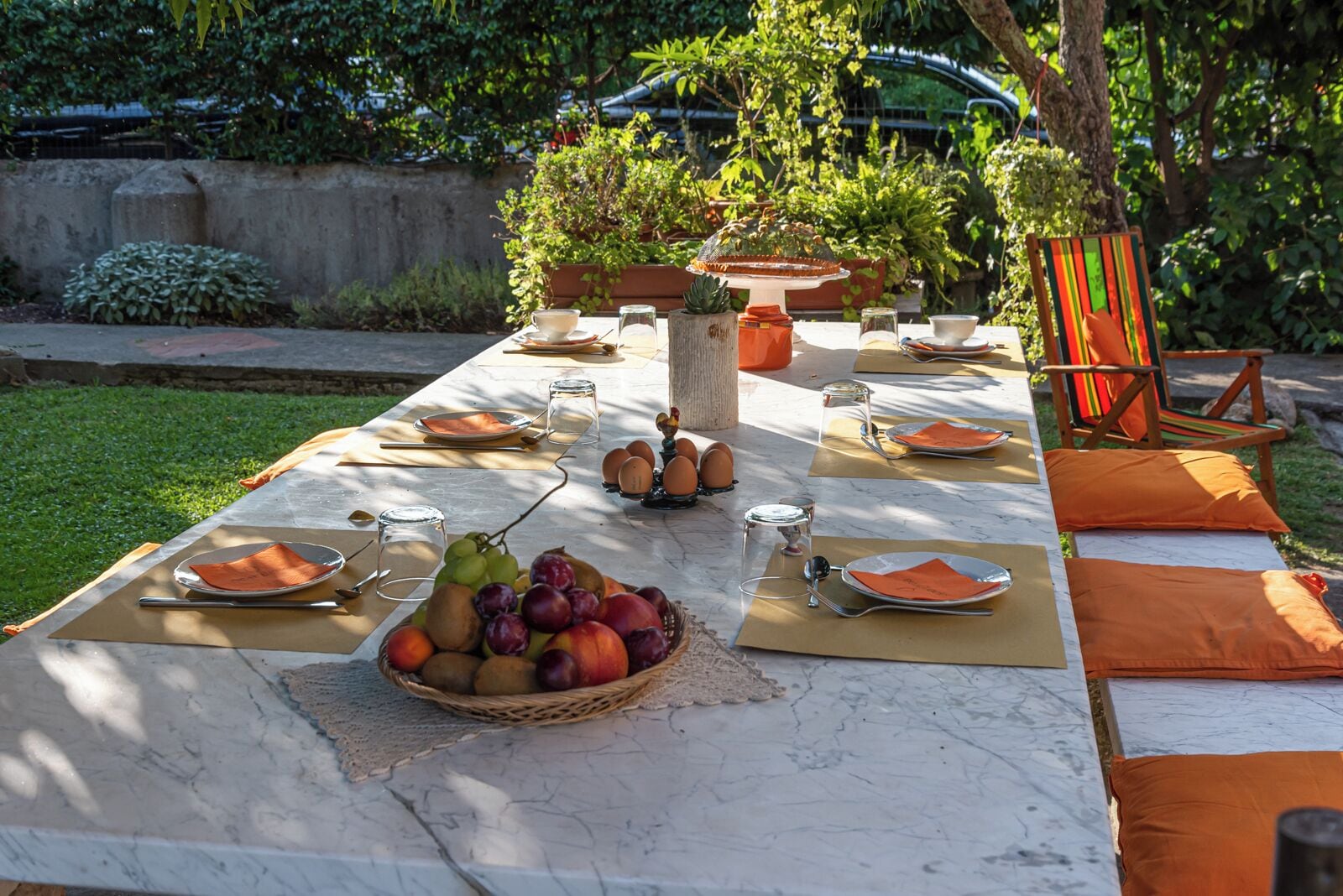 Una mesa de comedor exterior dispuesta con platos, cubiertos, vasos y una cesta de frutas.