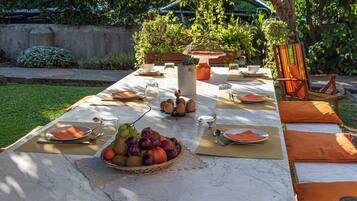 Una mesa de comedor exterior dispuesta con platos, cubiertos, vasos y una cesta de frutas.