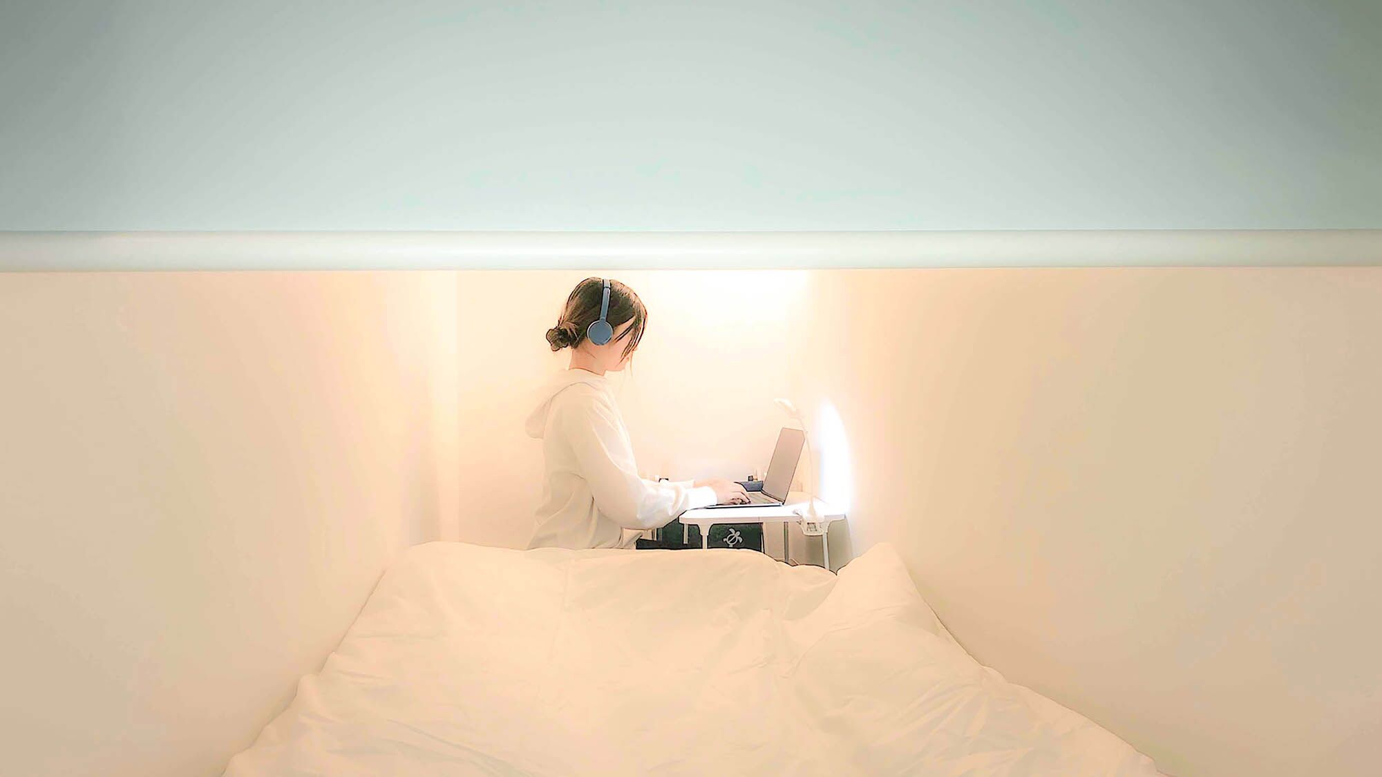 A person sitting at a desk in a room with a bed.