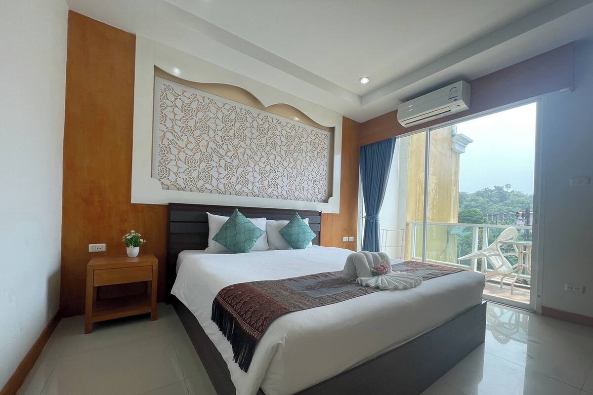 A modern hotel room with a large bed, a decorative wall panel, a small bedside table with a plant, and a balcony with a view.