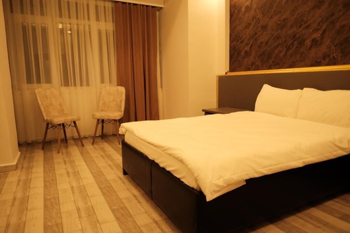 Ein Hotelzimmer mit einem großen Bett, zwei Sesseln, einem Nachttisch und einem Fenster mit durchsichtigen Vorhängen.