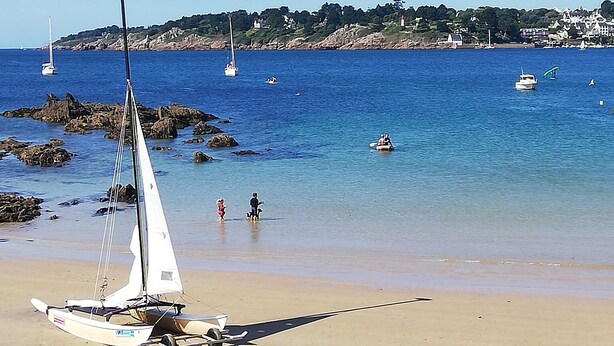 A beach with a sailboat, rocky shoreline, and boats in the water.