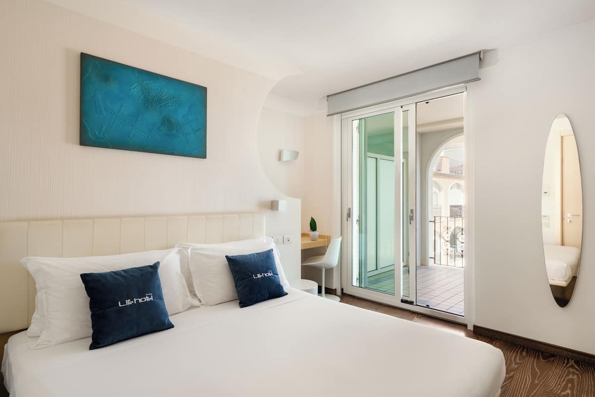 Habitación de hotel con cama, dos almohadas, un cuadro, una puerta corredera de vidrio, una silla y una mesita.