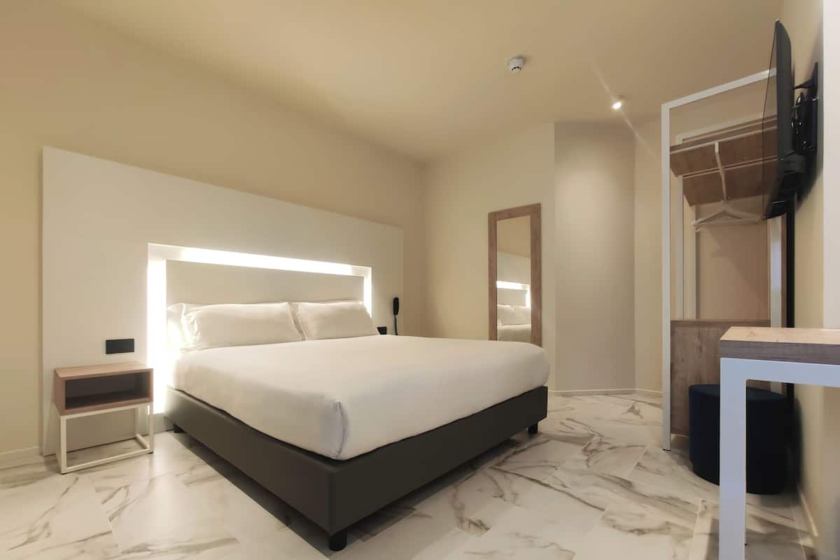 Una habitación de hotel moderna con una cama grande, una mesita de noche, un espejo, un televisor montado en la pared y una pequeña mesa.