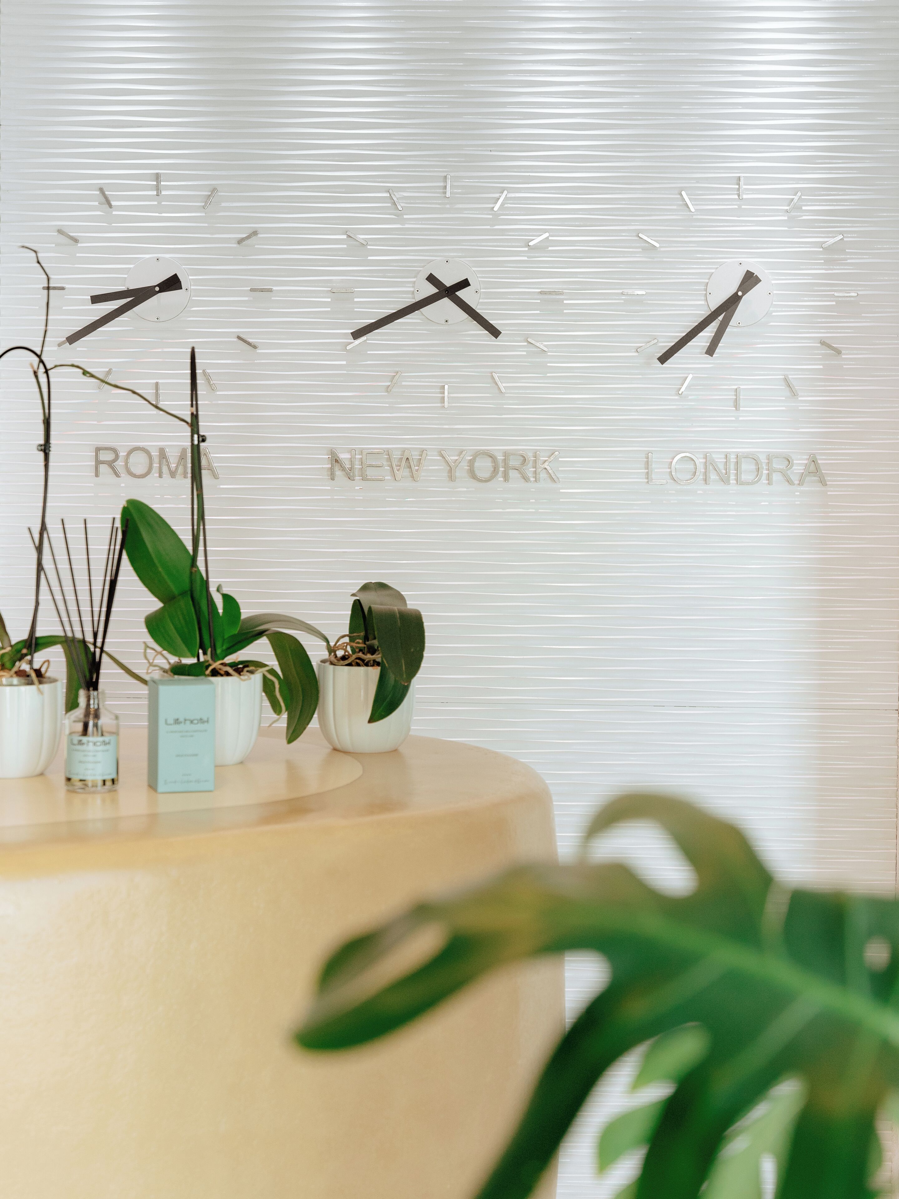 A reception desk with potted plants and a wall clock displaying 'ROME', 'NEW YORK', and 'LONDA'.