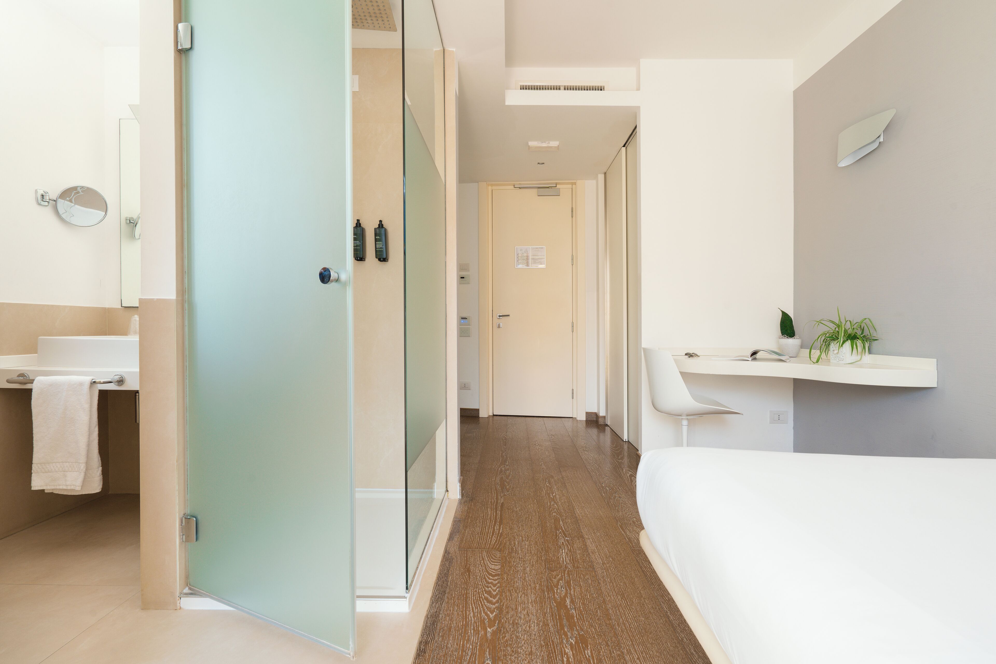 A modern hotel room with a wooden floor, a white bed, a bathroom with a mirror, and a shelf with plants.