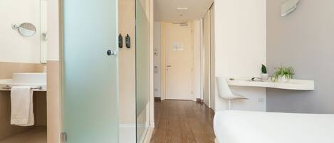 A modern hotel room with a wooden floor, a white bed, a bathroom with a mirror, and a shelf with plants.