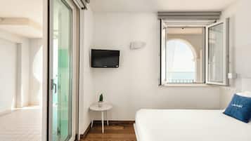 Habitación de hotel con una cama, un televisor montado en la pared, una mesita con una planta y una ventana con un persiano abierto.