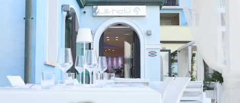 Un restaurante con terraza, un edificio azul al fondo y una pizza en primer plano.