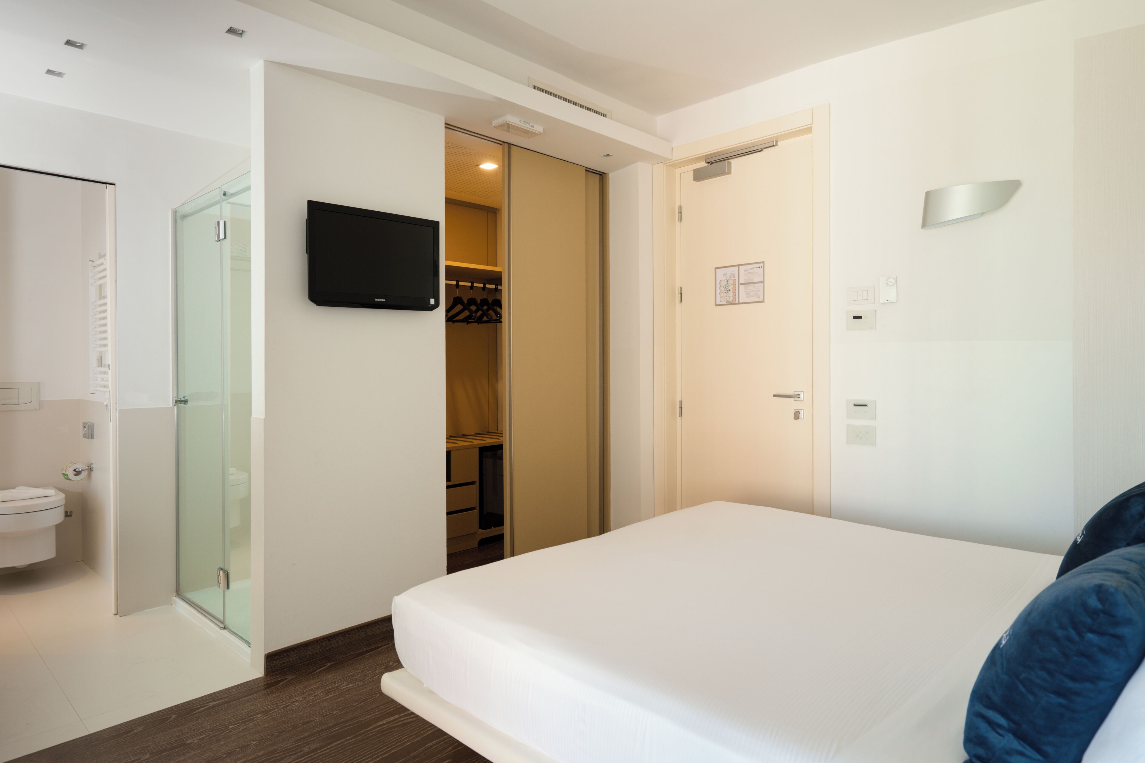 Habitación de hotel con cama, televisor montado en la pared, cabina de ducha de vidrio y armario.