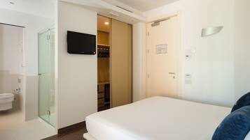 Habitación de hotel con cama, televisor montado en la pared, cabina de ducha de vidrio y armario.