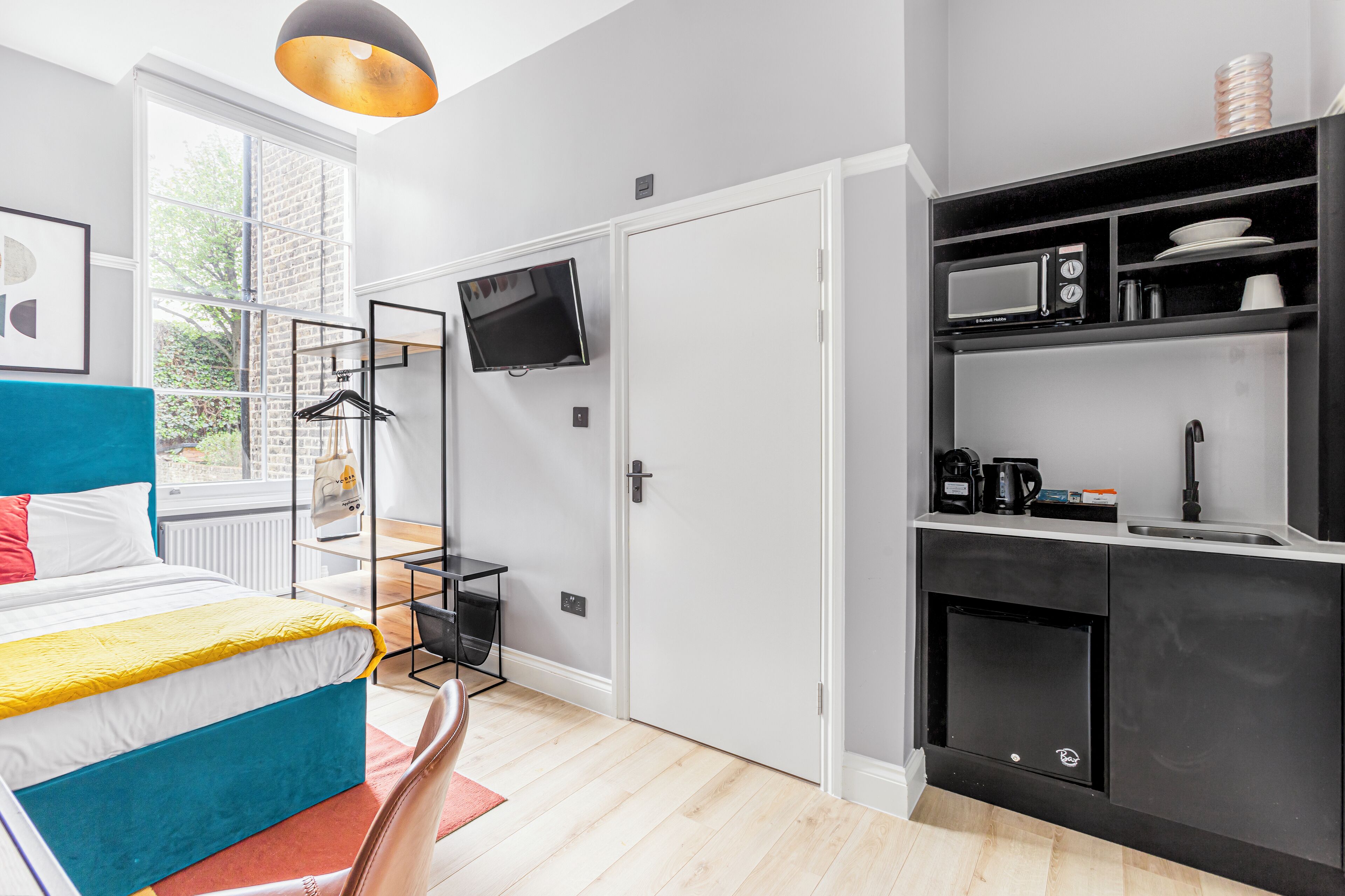 A modern room with a bed, a TV, a kitchenette, and a window with a view of greenery.