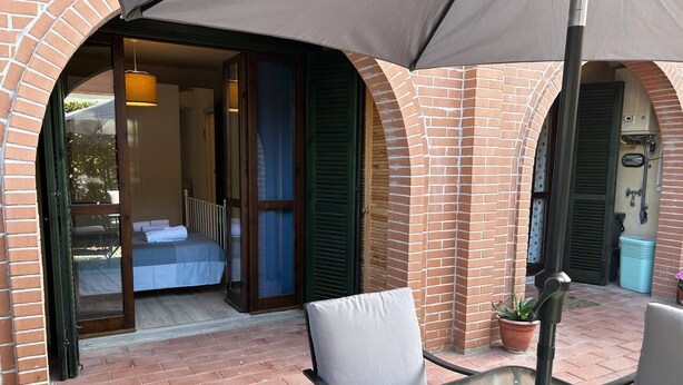 A patio area with a glass table and chairs, a brick wall, and a sliding glass door leading to a room with a bed. - Tea House. Apartment surrounded by greenery with garden, near the center of Lucca (Lucca)