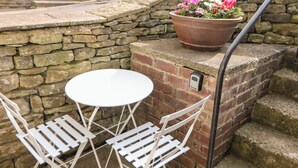 Un muro de piedra con una mesa y sillas blancas, una planta en maceta con flores y una barandilla de metal.