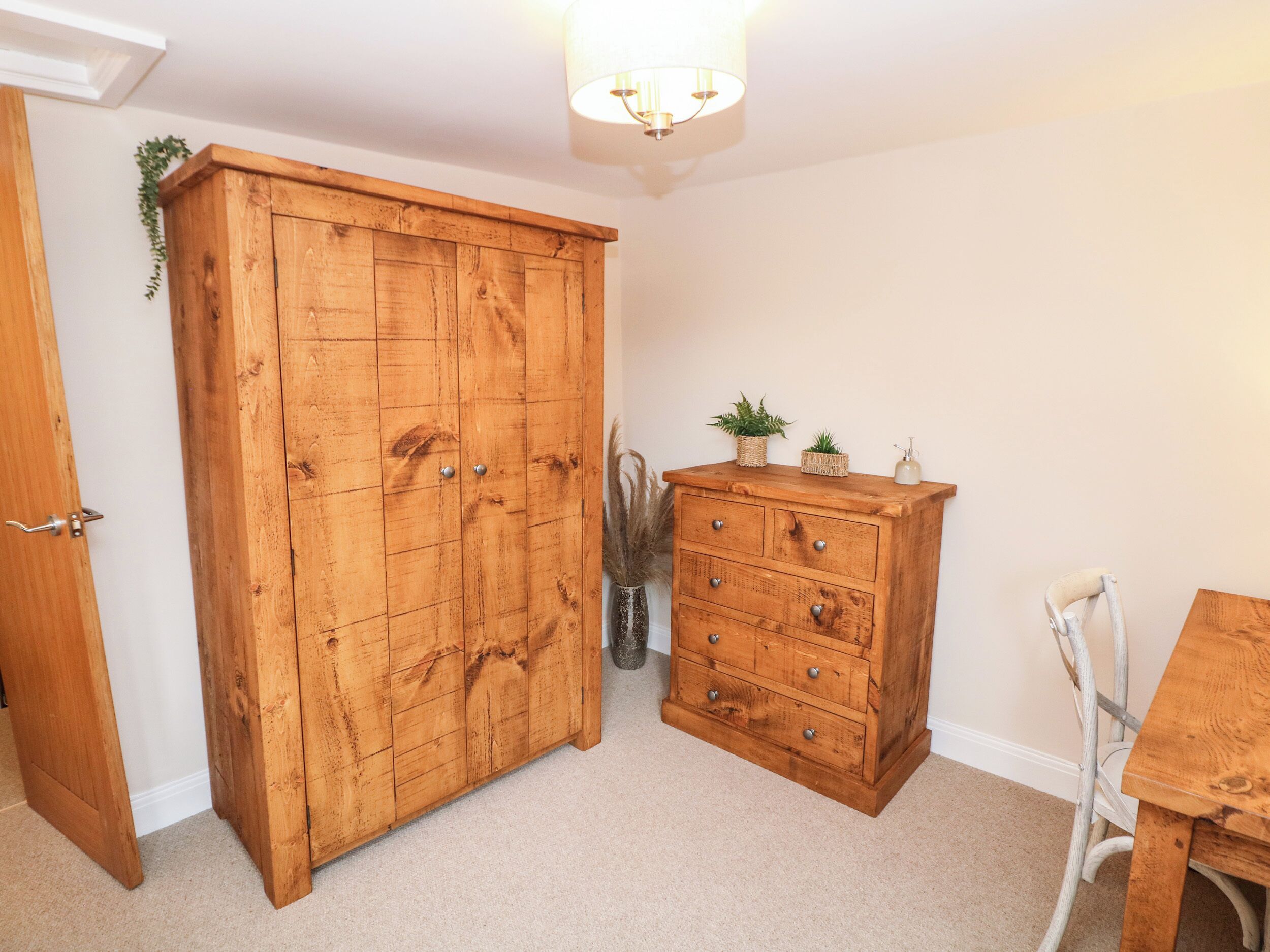 A room with a wooden wardrobe, a small wooden dresser with drawers, a white chair, and a table.