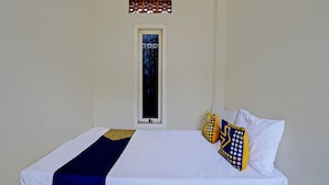 Una cama con una colcha blanca y azul, dos almohadas con diseños amarillos y azules, y una ventana pequeña con una celosía decorativa.