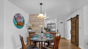 Un coin repas avec une table ronde, des chaises, un luminaire suspendu, un tableau et une cuisine en arrière-plan.