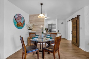 A dining area with a round table, chairs, a hanging light fixture, a painting, and a kitchen in the background.