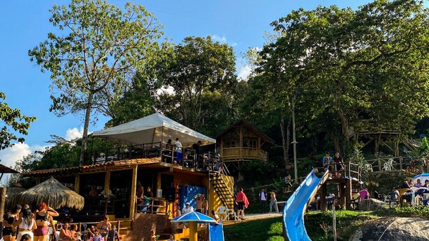 A pool area with a water slide, a wooden deck with people, and a building with a thatched roof. - Hostería El Sendero Cocorná (Cocorná)