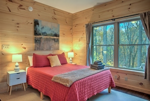 Un dormitorio de cabaña de madera con una cama, dos mesitas de noche, una ventana con cortinas y un cuadro en la pared.