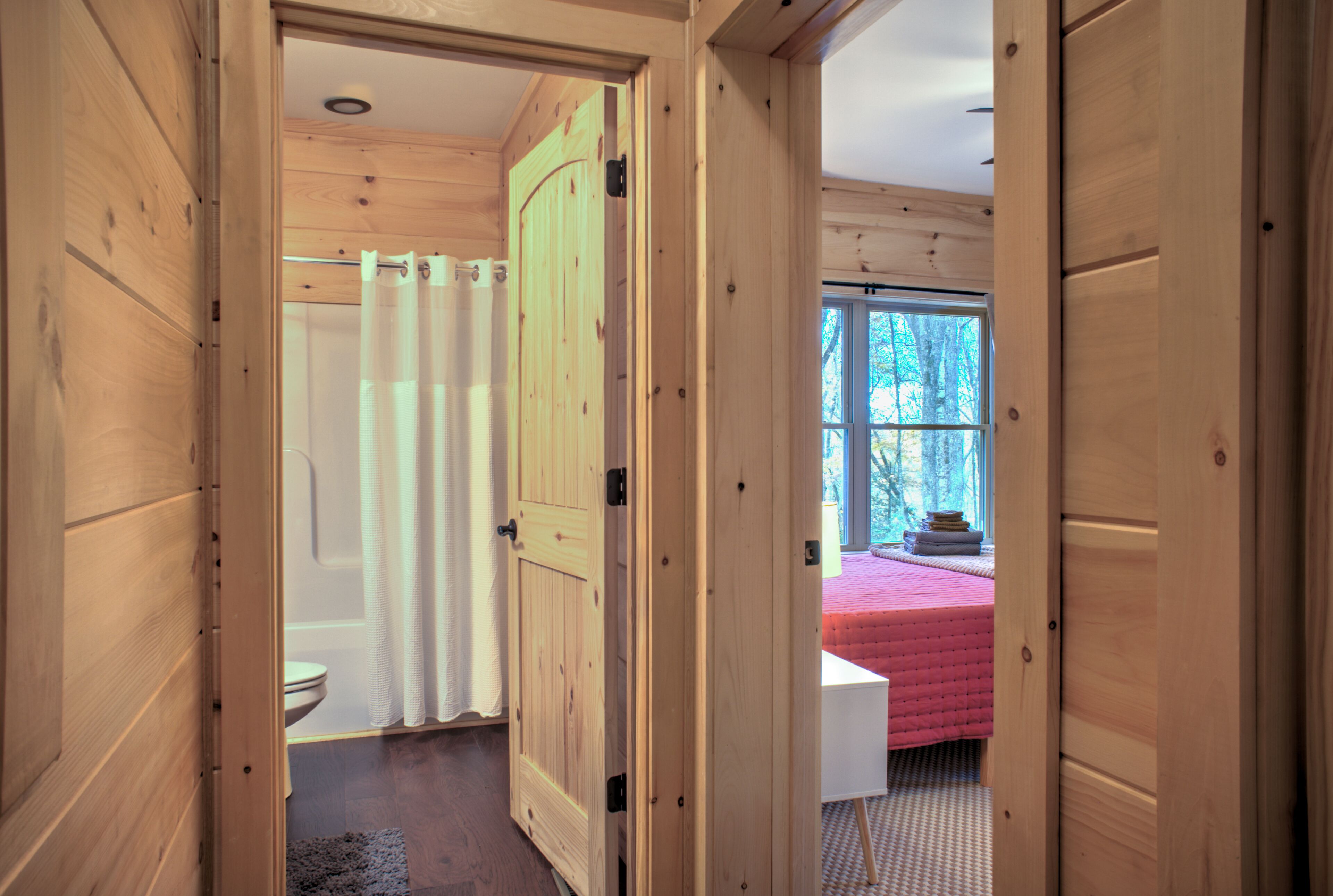 A wooden interior with a visible bathroom, a bed with a pink bedspread, and a window with a view of trees.
