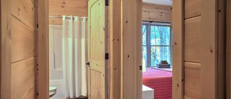 A wooden interior with a visible bathroom, a bed with a pink bedspread, and a window with a view of trees.