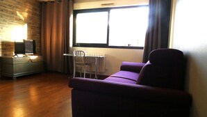 Olohuoneessa on violetti sohva, pieni pöytä ja tuoli, jalustalla oleva televisio sekä ikkuna, jossa on verhot.