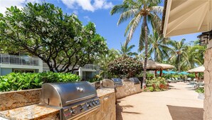 An outdoor grilling area with a stone wall, a grill, and a poolside lounge with umbrellas.