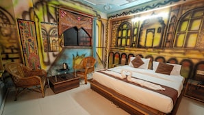 Un dormitorio con una cama grande, dos sillas de mimbre, una mesita y una pared con murales artísticos.
