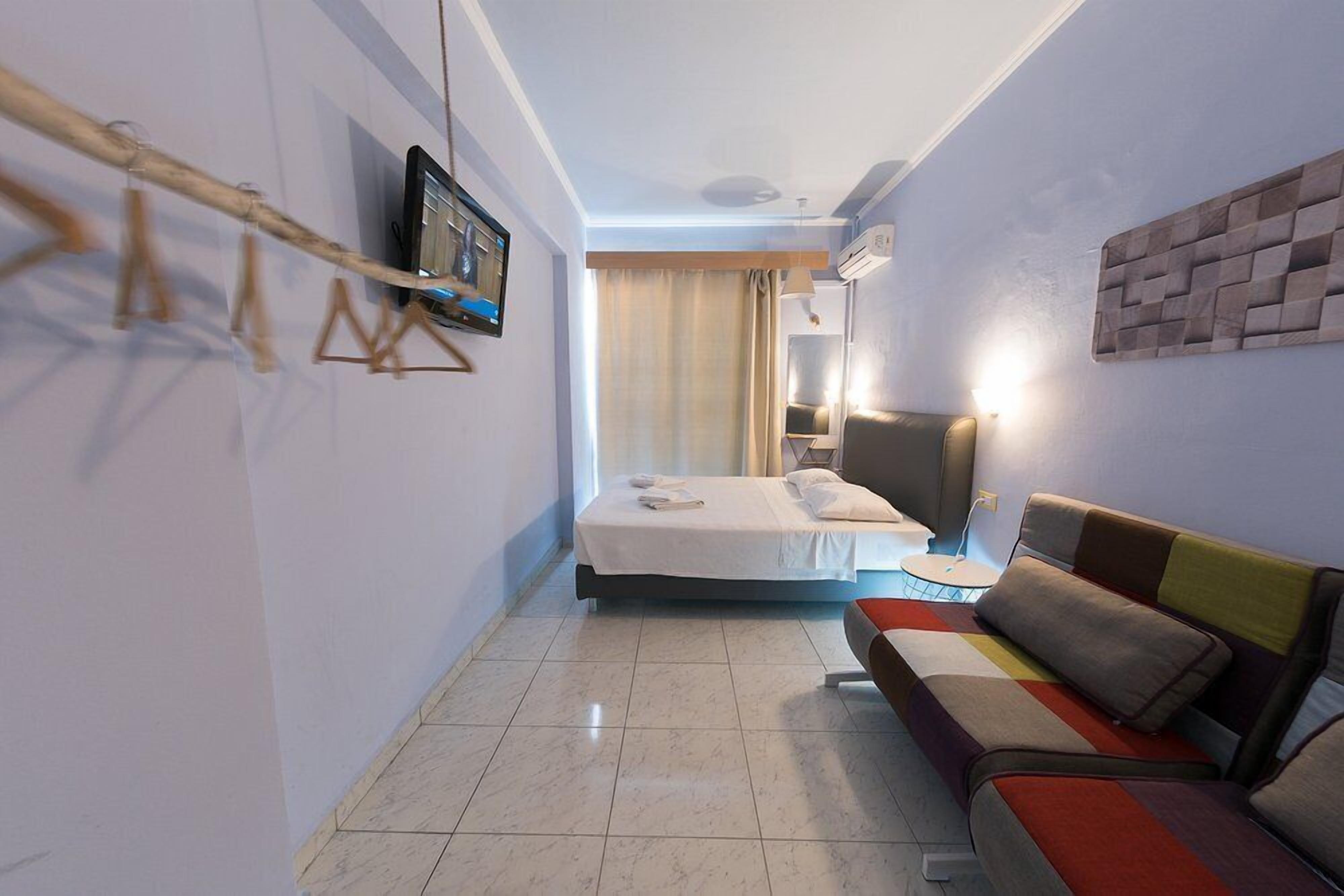 Una habitación de hotel con cama, sofá, televisión en la pared y ventana con cortinas.