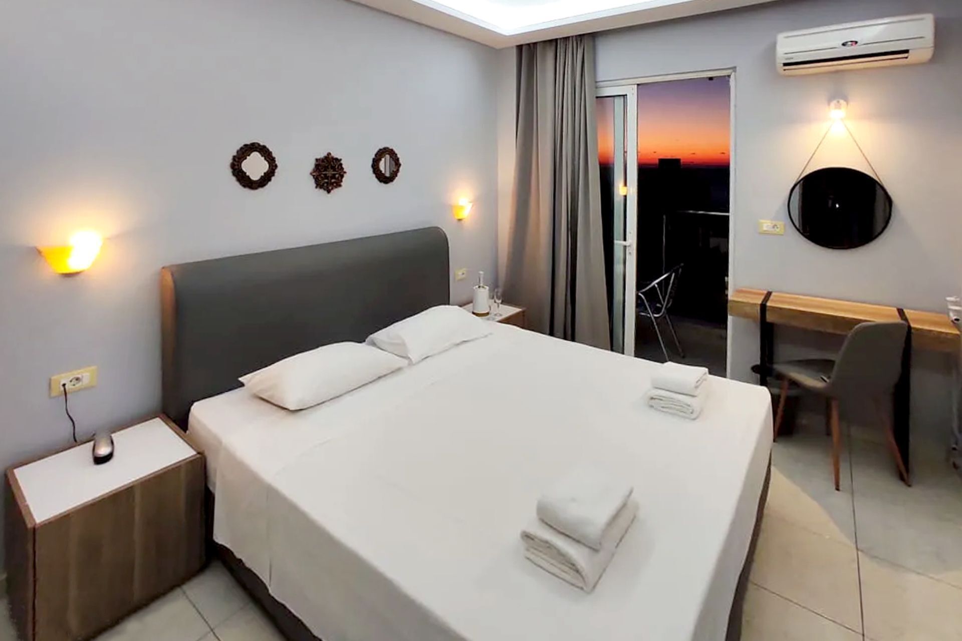 Habitación de hotel con cama, mesitas de noche, escritorio, silla, equipo de aire acondicionado y vistas al atardecer.
