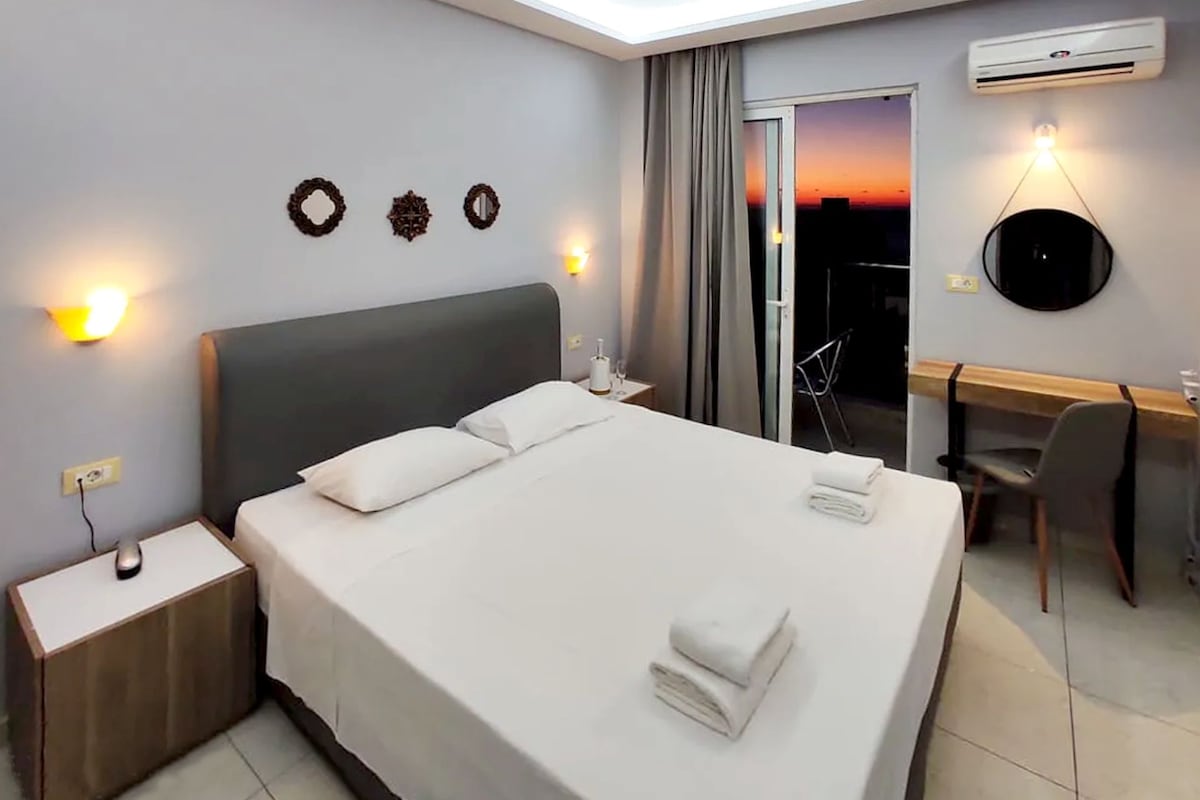 Una habitació d'hotel amb un llit, taules de nit, escriptori, cadira, aire condicionat i vista al capvespre.