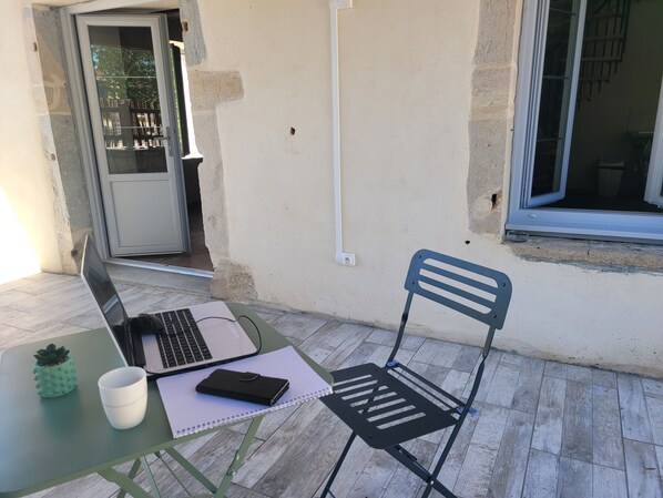 Une petite table d’extérieur avec un ordinateur portable, un carnet et une tasse, une chaise, et une porte donnant sur un balcon.