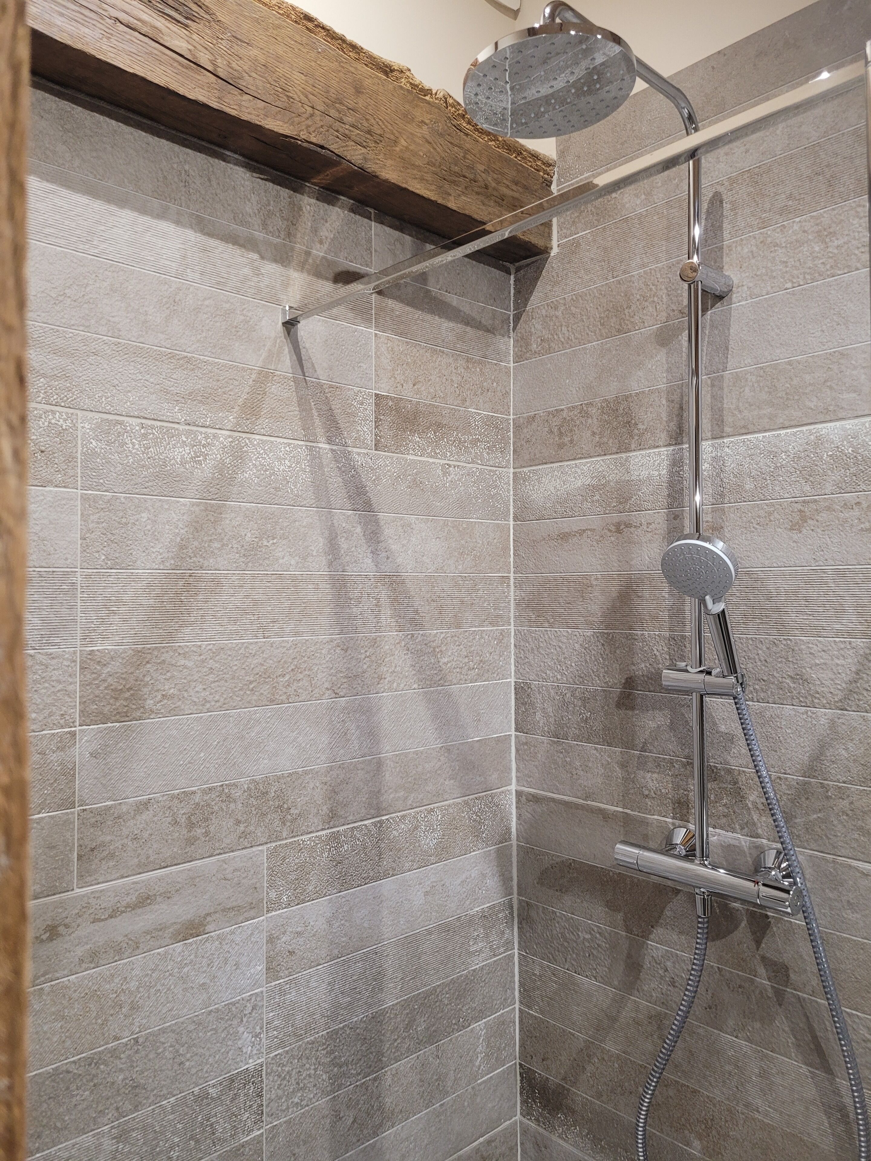 Un espace douche doté d’une pomme de douche au plafond et d’un pommeau de douche à main, de murs carrelés et d’une poutre en bois au plafond.