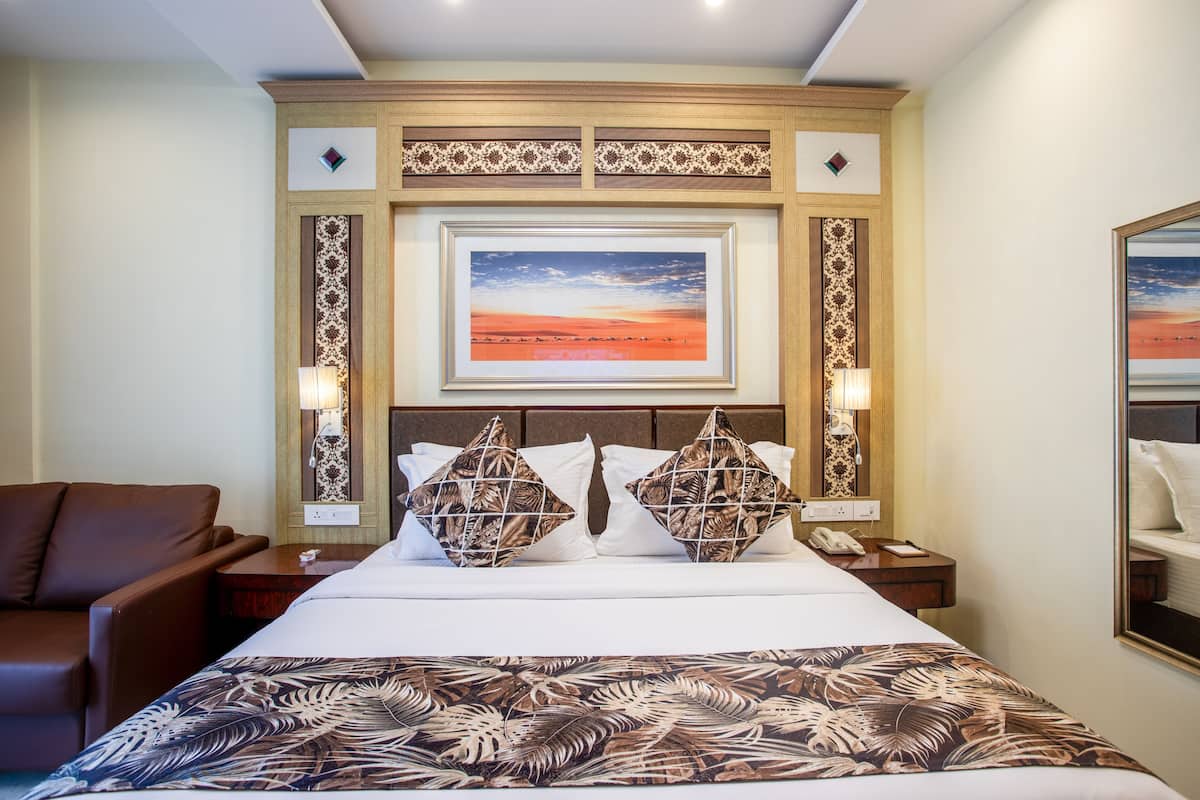 A hotel room with a large bed, two nightstands, a brown leather armchair, a framed sunset painting above the bed, and a patterned throw blanket.