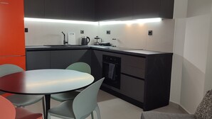 Een compacte keuken met een modern design, met een zwart aanrecht, een spoelbak en een waterkoker. Er is een klein zithoekje met een ronde tafel en twee stoelen.