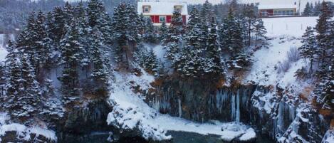 Un paysage hivernal avec une maison entourée d’arbres couverts de neige et une cascade gelée.
