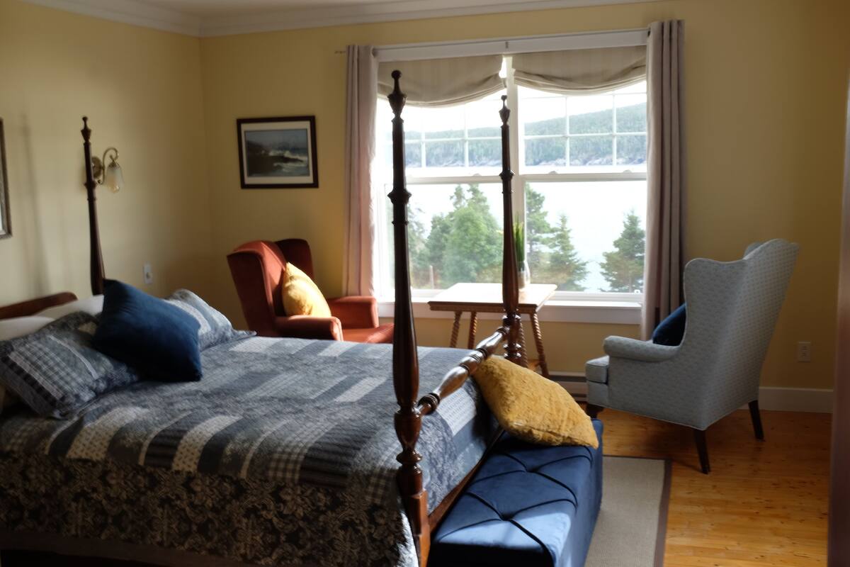 A bedroom with a four-poster bed, a chair, and a view of a lake through a window.