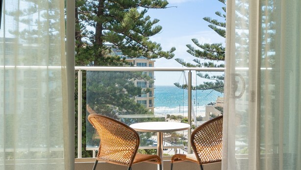 A balcony with wicker chairs and a small table, overlooking a coastal area with buildings and trees.