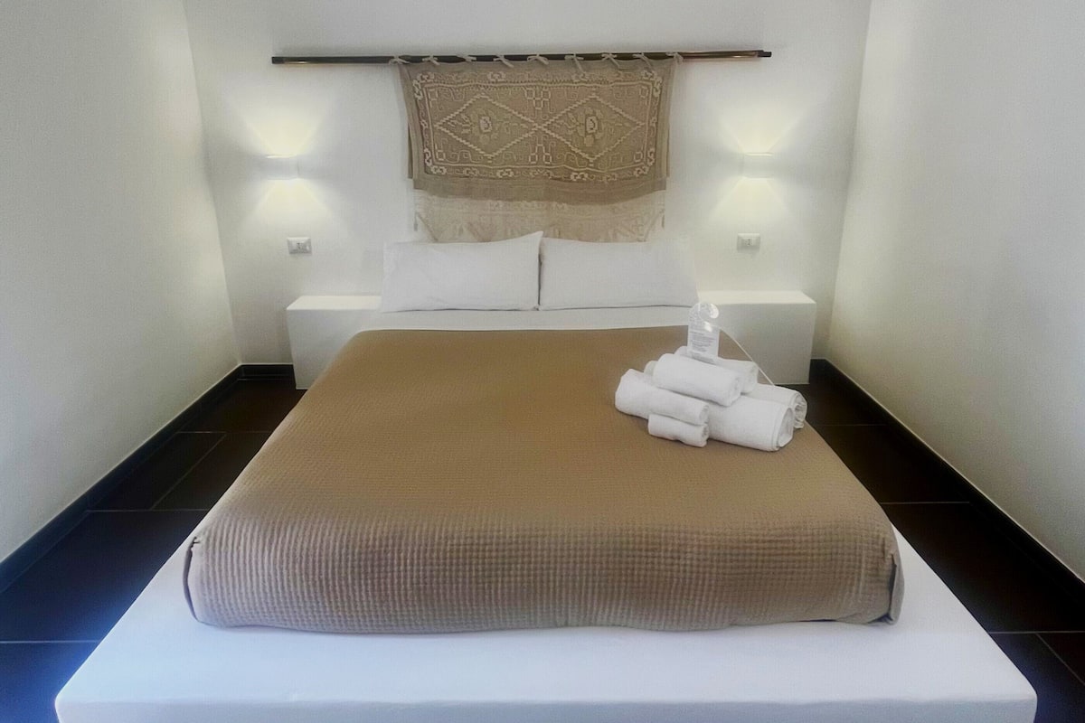 A modern hotel room with a large bed, a headboard with a decorative panel, two bedside tables with lamps, and a neatly folded towel on the bed.