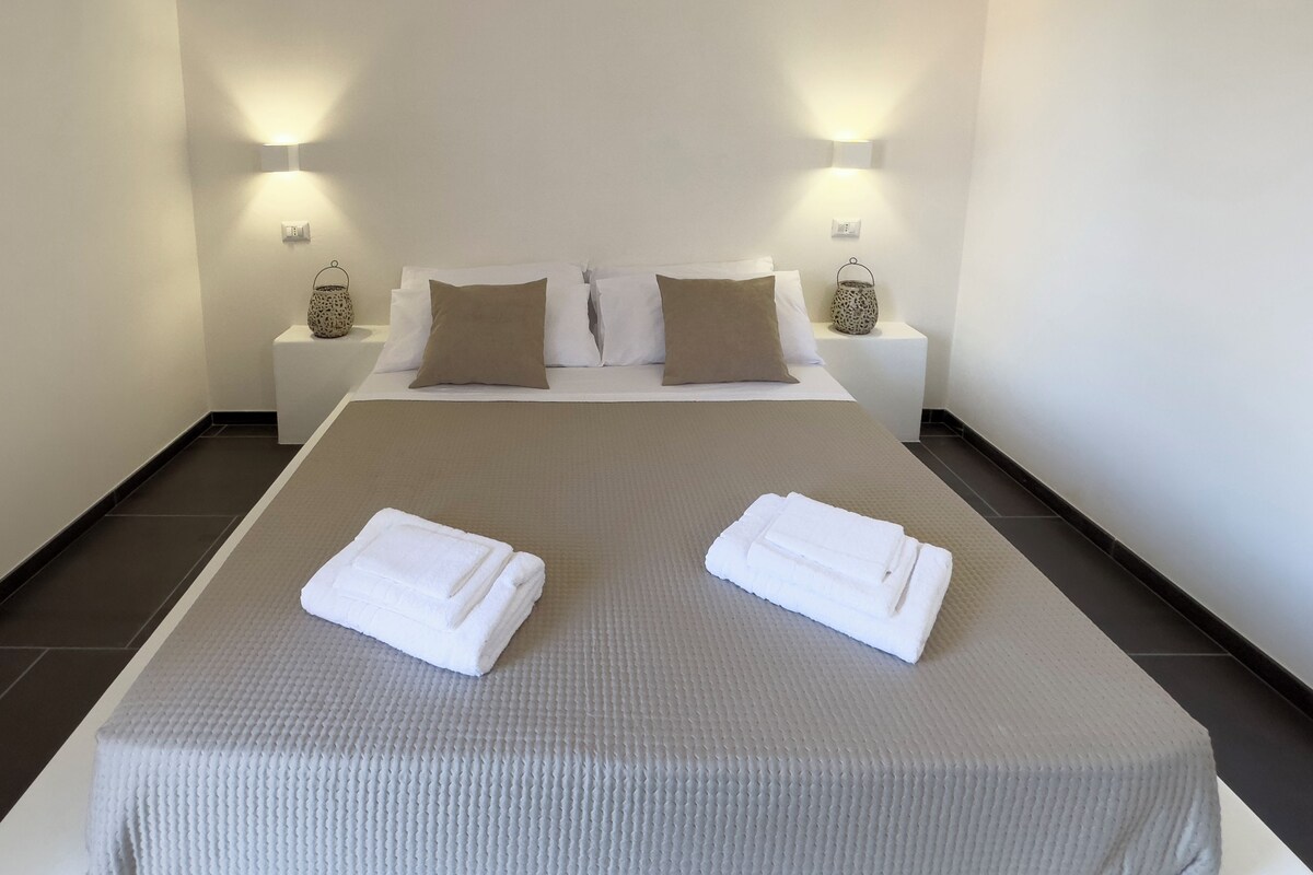 Un lit bien fait, avec deux serviettes blanches pliées, une couvre-lit beige et deux tables de chevet ornées de lampes et d’objets décoratifs.