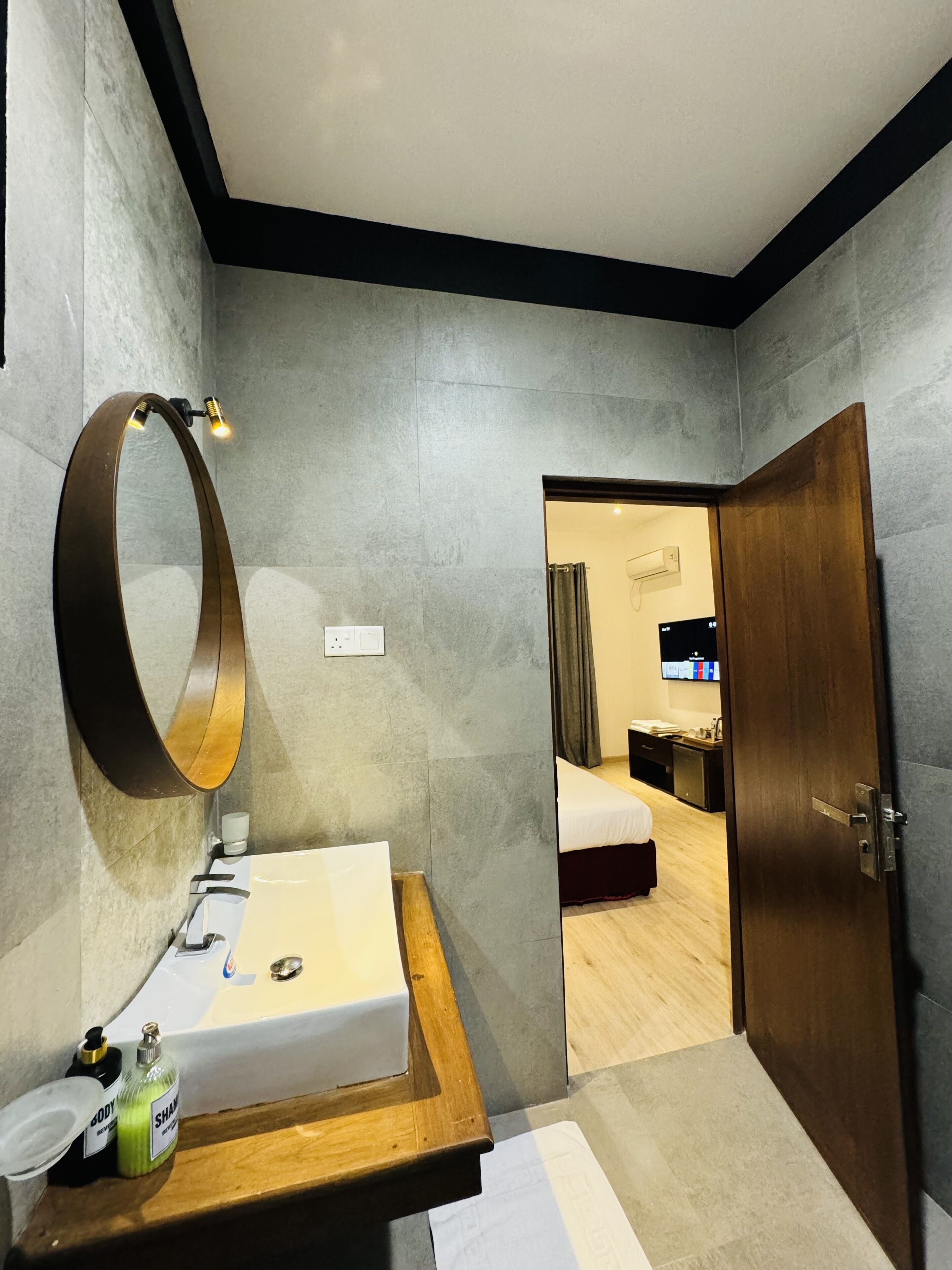 Un baño moderno con tocador de madera, un espejo grande y un lavamanos. Se ve una puerta que da a un dormitorio con una cama y un televisor.
