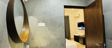 Un baño moderno con tocador de madera, un espejo grande y un lavamanos. Se ve una puerta que da a un dormitorio con una cama y un televisor.