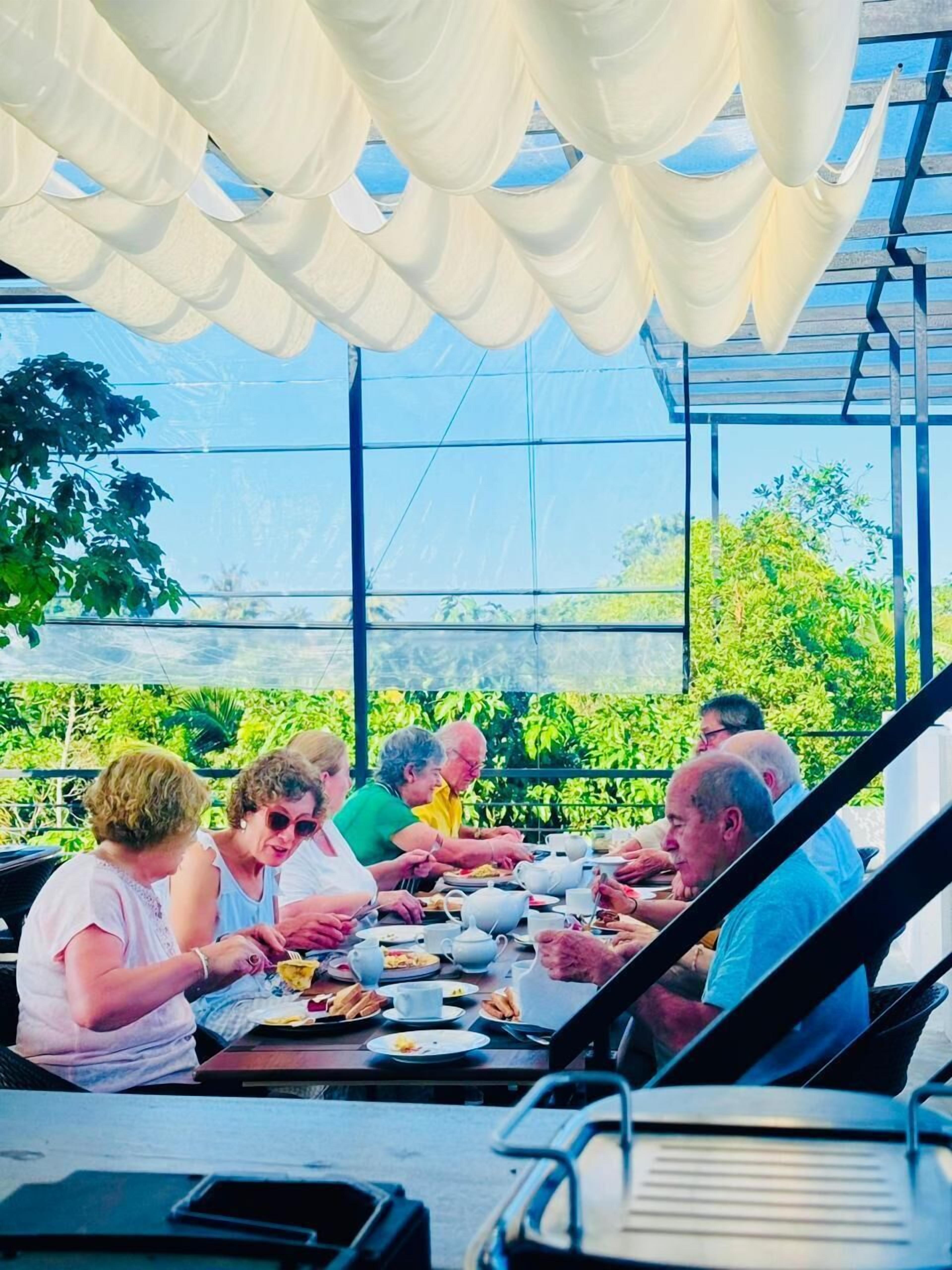 Un groupe de personnes est assis à une table en plein air, à l’abri sous une pergola, et profite de la vue sur la verdure.